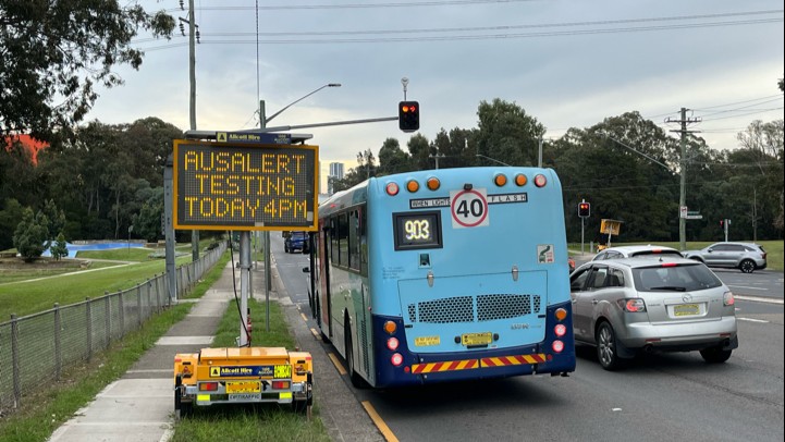 Photo of a temporary roadside sign that says AusAlert testing today 4 pm. On the road right next to it is the back of a blue bus and a silver car.