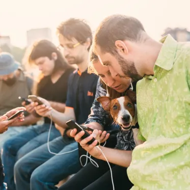 Group of people in a public space, looking at their phones. Focus is on two people looking at one phone with a dog in their lap.