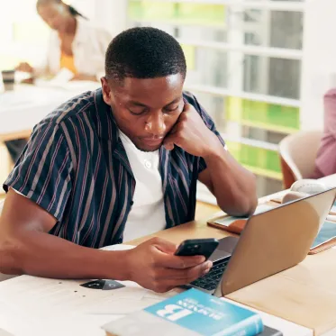 Student sitting at a desk in front of a laptop and looking at their phone.