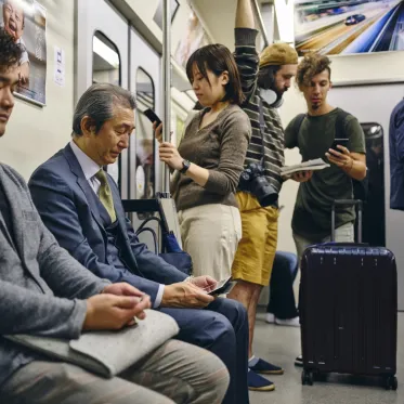 People standing and sitting on a train, looking at their phones. Some people are casually dressed with luggage, some are dressed in suits.