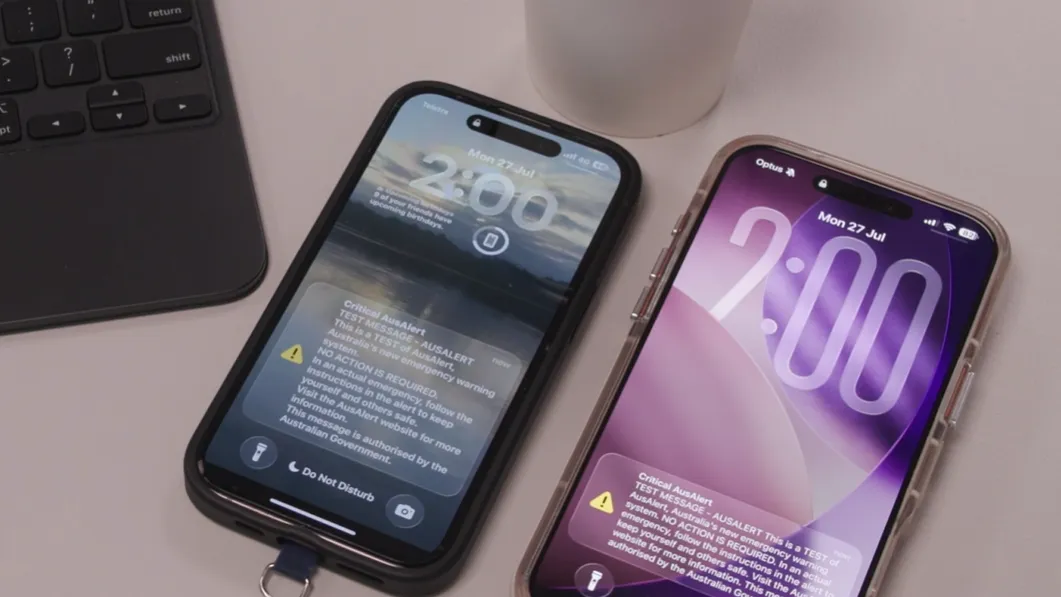 Photo of 2 smartphones on a desk, both showing 2pm and the AusAlert national test message on screen.