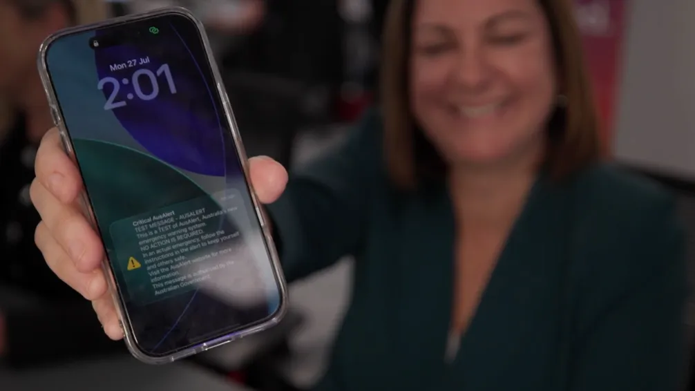 Photo of Minister McBain smiling, holding her phone forwards to the camera with the AusAlert national test on screen.